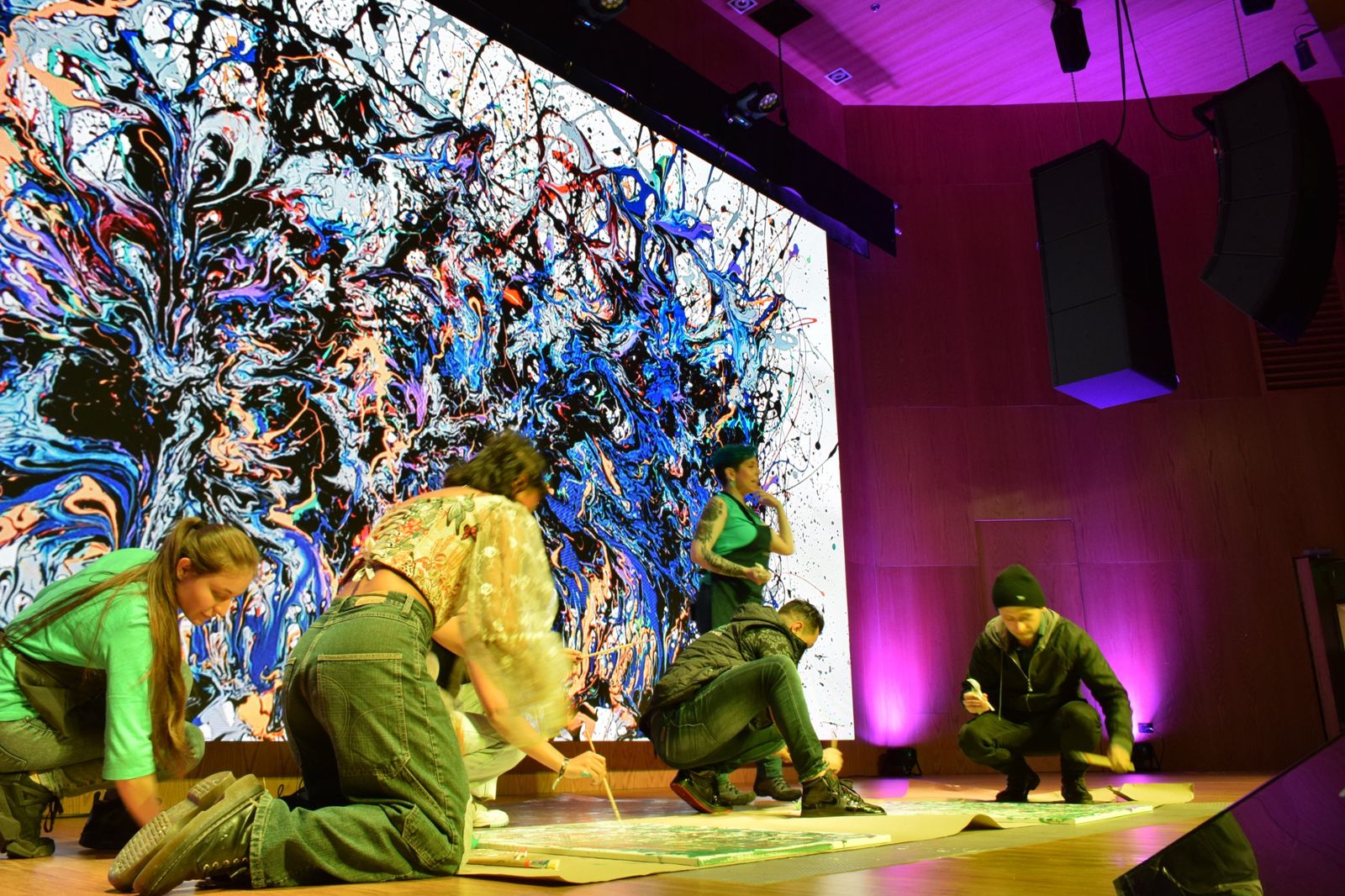 Participantes pintan en el escenario de un evento corporativo mientras la obra colectiva se proyecta en una pantalla gigante